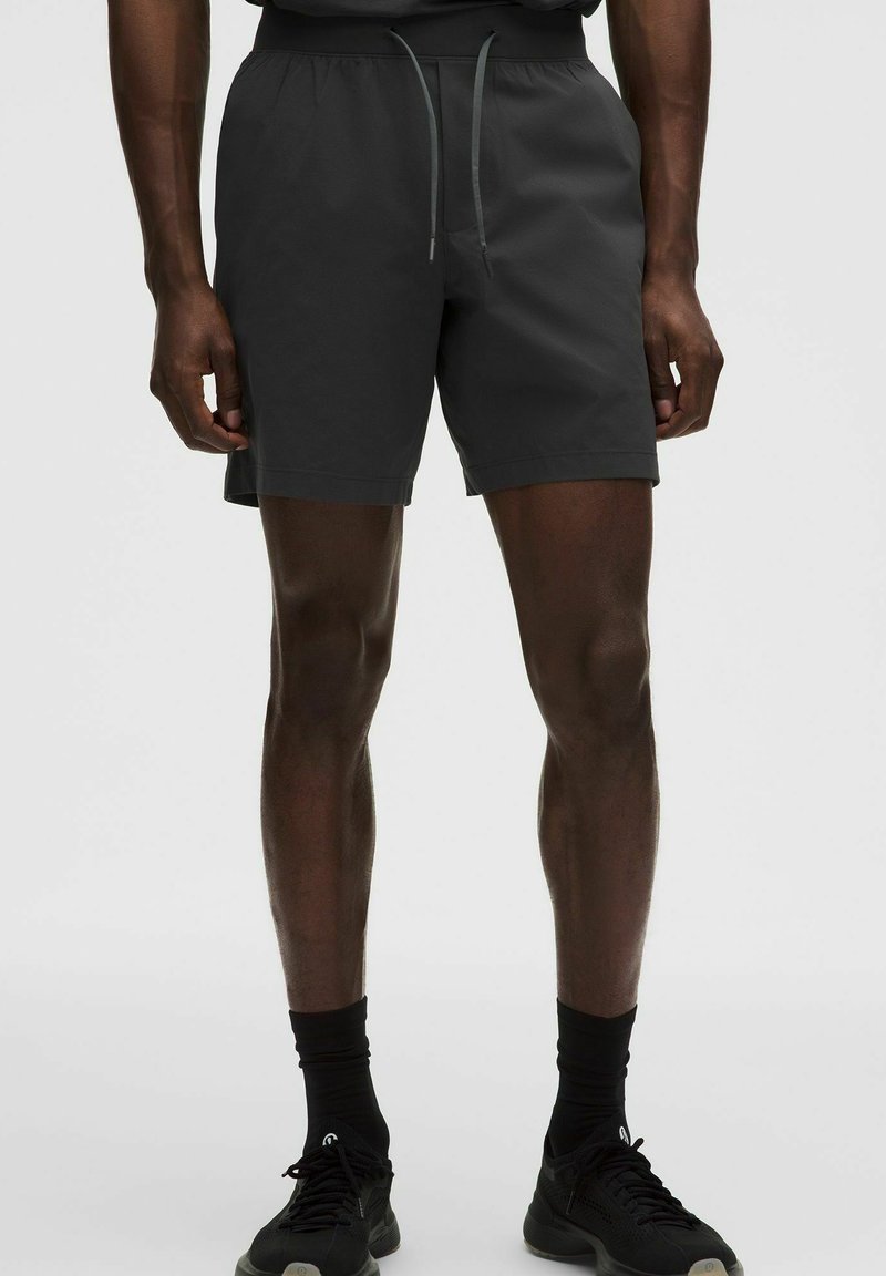 Mand iført sorte shorts med snøre, sorte sokker og sorte træningssko, stående foran en ensfarvet baggrund.