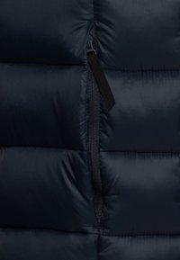 Närbild av en delvis stängd svart dragkedja på mörkblått vadderat quiltat tyg, som visar struktur och sömnadsdetaljer.