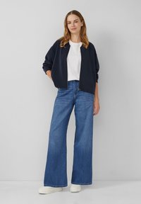 Temno modra bomber jakna, bela majica in široke modre kavbojke. Model nosi bele platforme, z dolgimi svetlo rjavimi lasmi v sproščenih valovih.
