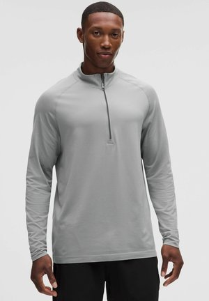 Man draagt een lichtgrijze pullover met lange mouwen en een kwart rits aan de voorkant, staand tegen een effen achtergrond.