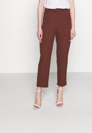 Frau trägt hochtaillierte, gerade geschnittene braune Hosen und weiße Sandalen mit Knöchelriemen und Absatz, steht vor einem schlichten hellen Hintergrund.