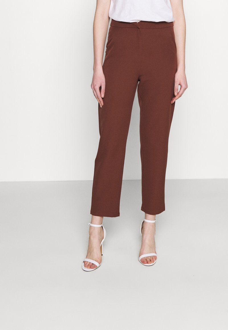 Femme portant un pantalon marron taille haute à jambes droites et des sandales blanches à talons avec bride à la cheville, debout devant un fond clair uni.