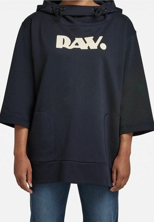 Personne portant un sweat à capuche noir ample avec des manches trois-quarts et des poches avant, avec le mot "DAY." en lettres crème en gras sur la poitrine.