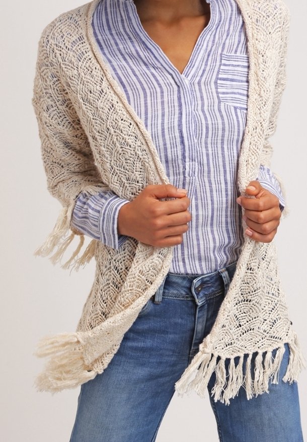 Gehaakte cardigan in crème, met een kantpatroon en franjes aan de onderrand. Draagt over een blauw-wit gestreepte blouse met kraag en denim jeans.