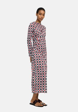 Kobieta ubrana w długą sukienkę o geometrycznym wzorze w kolorach czarnym, białym i czerwonym, do której dobrano czarne klapki ze srebrnymi pierścieniami.