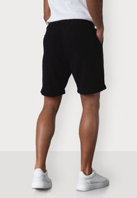 Schwarze Shorts aus einem weichen Material, mit einem klassischen Design, umgeschlagenen Säumen und Gesäßtaschen. Getragen mit weißen Sneakers.