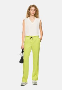Linen v-neck sleeveless top in white, paired with lightweight green pants featuring a drawstring and textured pattern. Holds a small black bag.