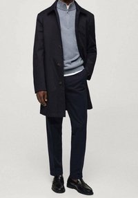 Marineblauer langer Mantel mit Knöpfen, hellgrauer Pullover mit Viertelreißverschluss, marineblaue Hose und schwarze Lederschuhe. Einfaches, modernes Design; keine Muster.