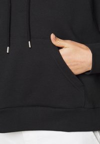 Sudadera negra con un bolsillo frontal, fabricada con un tejido texturizado, puños acanalados y cordones con terminales de metal. Pulgar visible en el bolsillo.