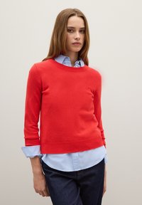 Maglione rosso a maglia con maniche lunghe, scollo rotondo e polsini a costine, indossato sopra una camicia azzurra a collare, abbinato a jeans in denim scuro.