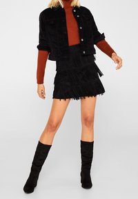 Svart cordjacka med två bröstfickor, kombinerad med en lager-på-lager svart fransad minikjol och höga svarta tygstövlar.