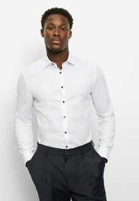 Weißes Langarmshirt mit Button-Down-Kragen und dunklen Knöpfen. Getragen mit dunkler Hose, glatter Textur und schmalem Schnitt.