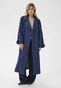 Dolga denim plašč v temno modri barvi, s širokim ovratnikom, dvojnimi gumbi in zavihanimi rokavi. Obučen je čez bež top.