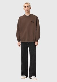 Sudadera marrón con cuello redondo, con un pequeño logo negro. Combinada con vaqueros negros holgados y zapatillas blancas.