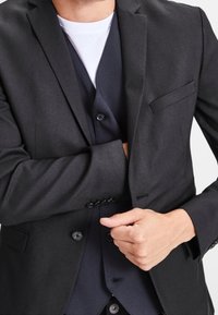 Czarny żakiet o teksturowanym wykończeniu, z pojedynczą kieszenią na piersi, zapinany na guziki, noszony na ciemnej kamizelce i białej koszuli.