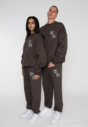 Twee modellen in bijpassende bruine sweatsuits met witte sneakers, staand tegen een eenvoudige witte achtergrond.