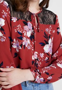 Röd blommig blus med en svart spetskrage. Har en knytning vid halsen och långa ärmar, med rosa och marinblå blommönster.