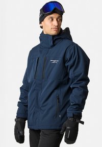 Navy blue weatherproof jacket with a high collar, front zip pockets, and black trim. Paired with black gloves and goggles.