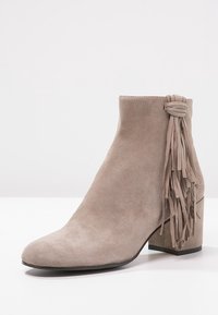Bota de tobillo de ante beige con un tacón grueso y un acento decorativo de flecos en el lateral. Textura suave y diseño de punta redonda.