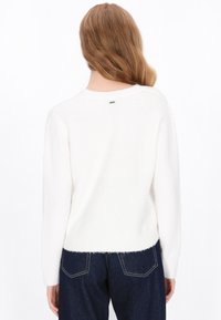 Hvid, fluffy sweater med lange ærmer, rund hals og en lille logoaccent på ryggen. Sammen med mørk denimjeans.