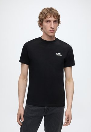 Ung mand med lyst krøllet hår iført en sort Karl Lagerfeld T-shirt og mørke jeans, stående mod en ensfarvet lys baggrund.