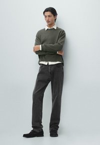 Grön stickad tröja lager-på-lager över en vit skjorta med krage, kombinerad med mörkgrå jeans och svarta skor. Enkel, modern design och avslappnad passform.