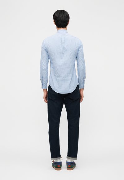 Világoskék hosszú ujjú ingben, sötét farmerben, fehér zokniban és kékeszöld vitorlás cipőben álló férfi, háttal látható, sima fehér háttér előtt.