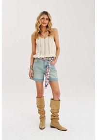 Pletený krémový tielko s čipkovým vzorom, svetlomodré džínsové šortky, vzorovaný šál previazaný v páse a béžové voľné vysoké čižmy po kolená.