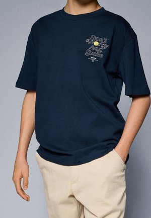Personne portant un T-shirt bleu marine avec le texte "Don't Worry And Smile" et un visage souriant sur la poitrine, associé à un pantalon beige.