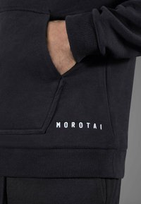 Felpa nera con tasca a marsupio, polsini a costine e logo "MOROTAI" bianco sull'orlo anteriore. Tessuto morbido e testurizzato.