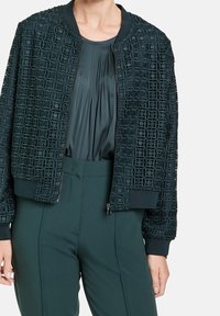 Chaqueta bomber verde con un patrón geométrico texturizado, puños y dobladillo acanalados. Combinada con una blusa plisada y pantalones verdes a medida.
