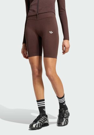 Persona que lleva puestos pantalones cortos de ciclismo marrones ajustados y chaqueta a juego, calcetines Adidas en blanco y negro y zapatillas deportivas negras con detalles plateados.