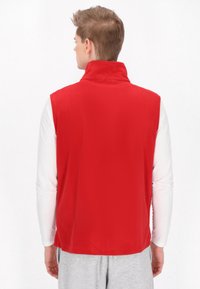 Roter ärmelloser Pullunder mit hohem Kragen, aus weichem Material. Getragen über einem weißen Langarmshirt, kombiniert mit grauen Hosen.