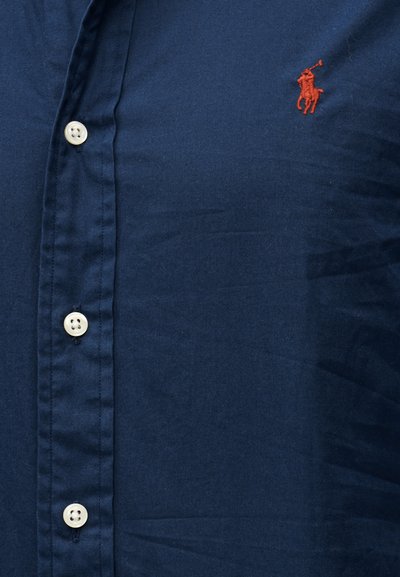 Tengerészkék gombos ing fehér gombokkal és egy kis piros hímezett póni játékos logóval a bal mellrészen.