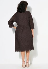 Dunkelbraunes Kleid aus Lochstickerei mit geometrischen Mustern und einem gewellten Saum. Dreiviertelärmel und eine taillierte Passform.
