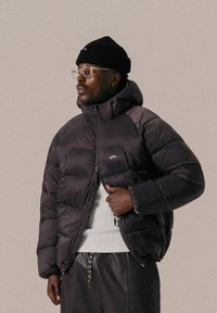 Doudoune noire avec capuche, sections matelassées bien visibles, fermeture éclair à l'avant et petit logo sur la poitrine, associée à un pull blanc.