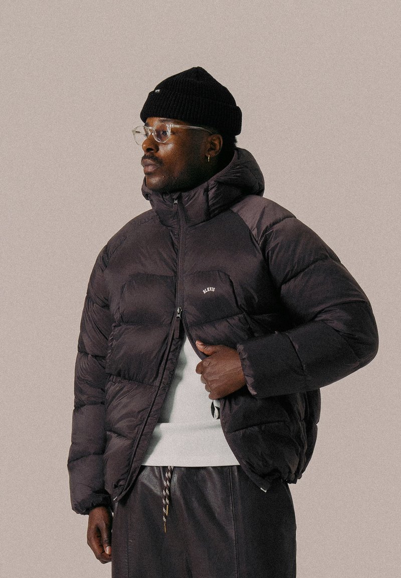 Doudoune noire avec capuche, sections matelassées bien visibles, fermeture éclair à l'avant et petit logo sur la poitrine, associée à un pull blanc.