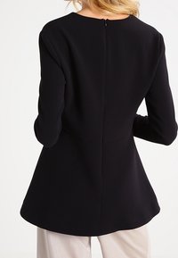 Maglietta nera a maniche lunghe con scollo rotondo, corpo aderente e orlo svasato. Presenta una cerniera nascosta sul retro e una texture di tessuto liscia.