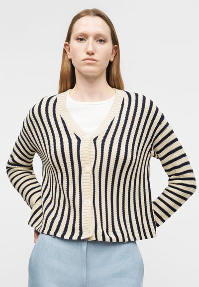 Mladá žena so svetlohnedými rovnými vlasmi má na sebe béžový a čierno pruhovaný kardigán cez bielu košeľu a svetlomodré nohavice.