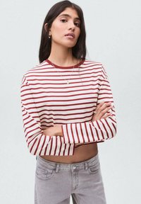 Röd och vit randig långärmad crop top med rund halsringning, kombinerad med ljusgrå jeans. Slät textur, avslappnad design.