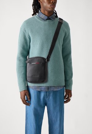 Black leather crossbody bag with textured surface, red accent, and adjustable strap, worn over a teal ribbed sweater and denim shirt.