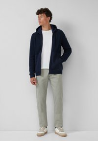 Navy zip-up hoodie met ribbeltextuur, gecombineerd met een wit t-shirt en lichtgrijze broek. Bevat een capuchon en zijzakken. Casual sneakers.