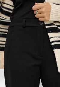Pantalon noir au design épuré, avec une fermeture à bouton, des poches latérales et une texture lisse, associé à un cardigan en tricot rayé.