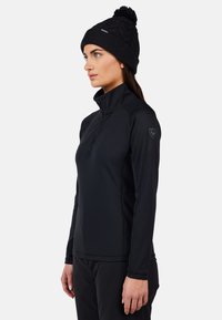 Fleece nero con zip e colletto alto, caratterizzato da un design aderente e polsini elastici. Indossato con un berretto di lana nero con pom-pom.