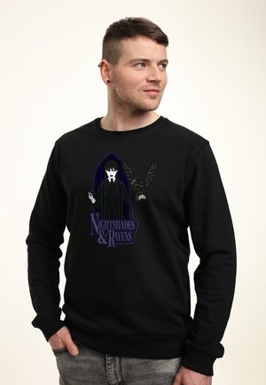Mężczyzna w czarnej bluzie z grafiką w stylu gotyckim przedstawiającą osobę w pasiaku i kruka, z podpisem "Nightshades & Ravens."