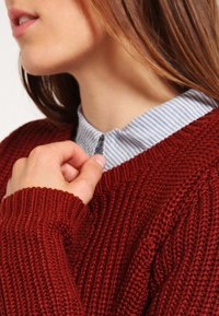 Maglione a coste bordeaux con scollo ampio, indossato sopra una camicia a righe azzurre con colletto, che presenta dettagli con bottoni.