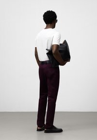 Camicia bianca a maniche corte, pantaloni viola, cintura nera e grande borsa di pelle nera. Sandali neri, capelli ricci e occhiali da sole. Vista da dietro.