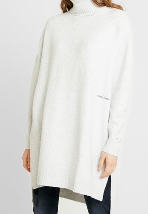 Vrouw draagt een oversized witte coltrui met geribbelde manchetten en zoom, gecombineerd met donkere jeans, tegen een effen achtergrond.