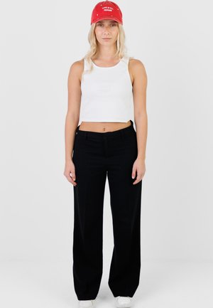 Vaaleahiuksinen nainen, jolla on punainen lippalakki, jossa lukee "LOVE IS ALL AROUND", valkoinen hihaton lyhyt toppi, mustat leveälahkeiset housut ja valkoiset kengät, seisoo valkoisen taustan edessä.