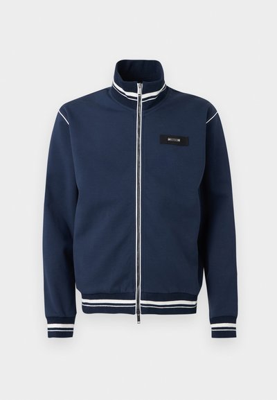 Veste zippée bleu marine avec des détails de bandes blanches sur le col, les poignets et l'ourlet, et un petit patch logo rectangulaire sur la poitrine gauche.